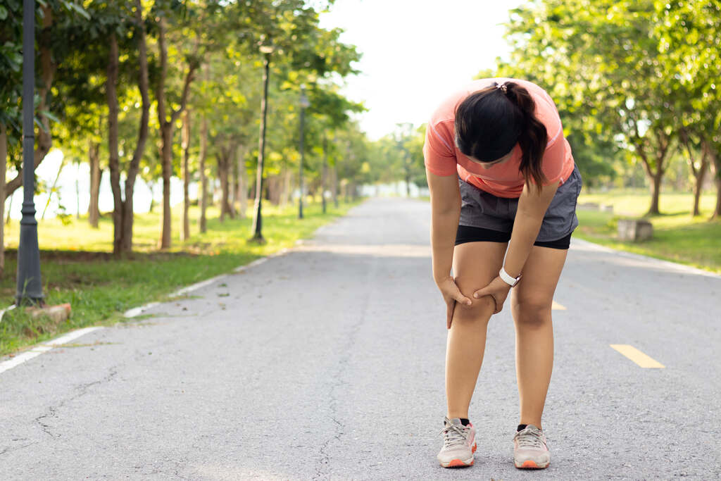 Uma mulher em roupas esportivas (camiseta rosa e shorts cinza) para no meio de uma pista de caminhada em um parque para segurar o joelho, indicando dor ou lesão. Ela está levemente curvada e o fundo mostra árvores e iluminação solar suave.
