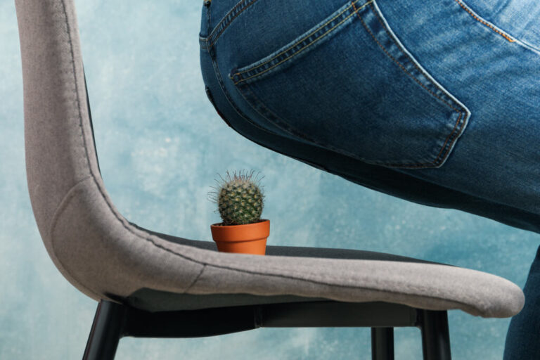 Uma pessoa vestindo calça jeans faz menção de se sentar em uma cadeira de escritório onde há um pequeno cacto em um vaso, simbolizando desconforto ao sentar.